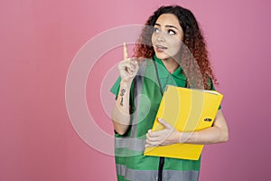 Curious, pretty woman posing indoors, looking up, pointing by finger, holding folder.