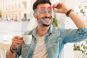 Curious and smiling casual man fixing sunglasses drinks coffee