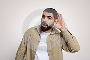 Curious young man with a beard, hand to ear, actively listening, expressing attention and interest