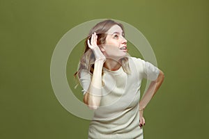 Curious young woman with hand near ear listening rumor, gossips on green background. Deafness concept. Hearing gesture