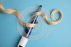 Curling iron with blonde hair lock on light blue background, flat lay