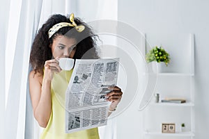 Curly african american woman reading newspaper and covering face with cup