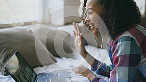 Curly african american young woman having video chat with friends using laptop camera while lying on bed