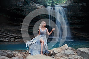 A curly blonde girl in a luxurious blue dress sits on white stones against the backdrop of a fabulous landscape. River