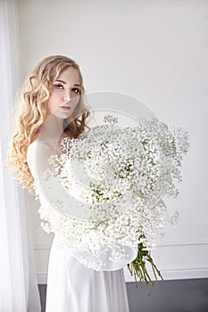 Curly blonde romantic look, beautiful eyes. White wildflowers in hands. Girl white light dress and curly hair, portrait of woman