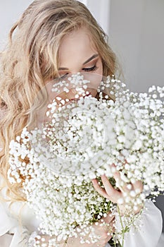 Curly blonde romantic look, beautiful eyes. White wildflowers in hands. Girl white light dress and curly hair, portrait of woman