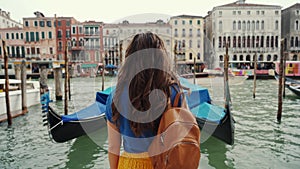 Curly brunette girl in blue t-shirt stops near gondolas, Grand Canal. Rear view