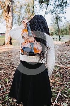 curly brunette woman holding a violin on her back, walking through the park road at sunset. Vertical