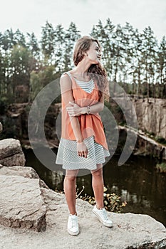 Curly dark-haired woman wearing striped baggy summer dress standing near lake