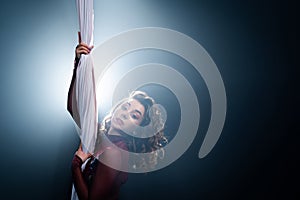 Curly female gymnast performs on a black background with rays. circus artist on white aerial silks