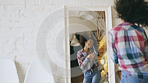 Curly funny african american girl dancing and singing with hair dryer in front of mirror at home