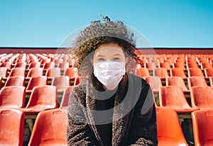 Curly girl in a surgical mask sitting in an empty stadium during epidemic disease Covid-19