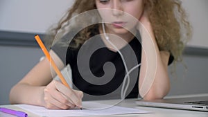 Curly girl teenager listening music in earphones and writing by pencil on paper sheet. School girl writing text by