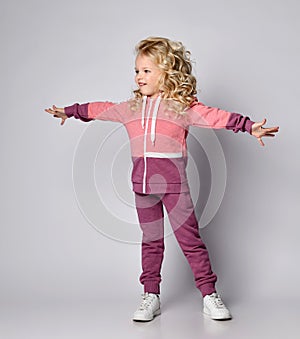 Curly hair blonde kid girl in modern fashion pink gray sportsuit stands with her arms outstretched wide spread
