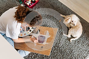 Curly hair smiling girl teenager in glasses in white t-shirt sitting on floor and painting on wooden desk with poodle dog at the