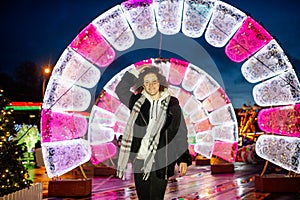 Curly hair smiling young woman in a coat outdoors with Christmas lights and decoration in the background