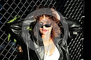 Curly hair woman in sunglasses and leather jacket posing on dark background