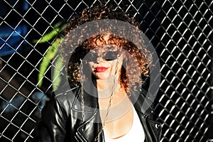 Curly hair woman in sunglasses and leather jacket posing on grid background