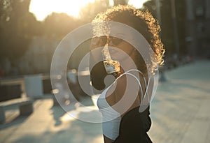 Curly hair woman in sunglasses and leather jacket posing outdoors at sunset.