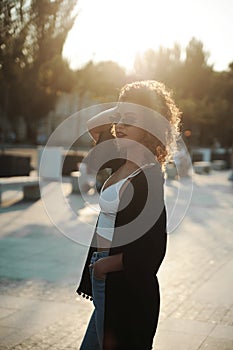 Curly hair woman in sunglasses and leather jacket posing outdoors at sunset.