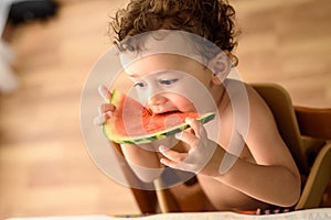 Curly-haired baby girl eats fruit sitting indoors at home in summer