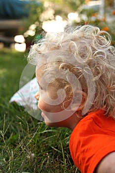 Curly haired, blond, smiling toddler outdoors on grass in garden.