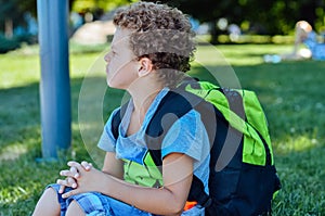 Curly-haired boy sits on the ground with a large green backpack closed his eyes and framed hands the boy is surprised