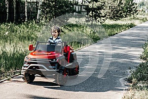 Curly-haired boy in a striped T-shirt rides a red big toy car driving on an asphalt path. day off, outdoor recreation