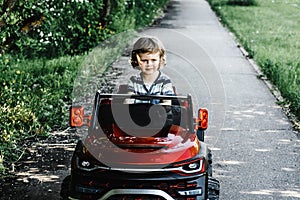 Curly-haired boy in a striped T-shirt rides a red big toy car driving on an asphalt path. day off, outdoor recreation