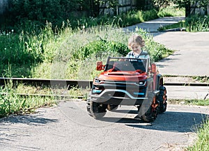 Curly-haired boy in a striped T-shirt rides a red big toy car driving on an asphalt path. day off, outdoor recreation