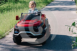 Curly-haired boy in a striped T-shirt rides a red big toy car driving on an asphalt path. day off, outdoor recreation