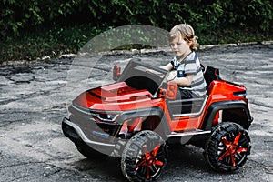 Curly-haired boy in a striped T-shirt rides a red big toy car driving on an asphalt path. day off, outdoor recreation