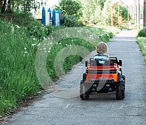 Curly-haired boy in a striped T-shirt rides a red big toy car driving on an asphalt path. day off, outdoor recreation