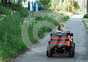 Curly-haired boy in a striped T-shirt rides a red big toy car driving on an asphalt path. day off, outdoor recreation