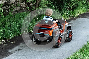 Curly-haired boy in a striped T-shirt rides a red big toy car driving on an asphalt path. day off, outdoor recreation