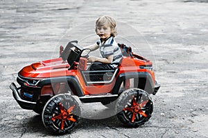 Curly-haired boy in a striped T-shirt rides a red big toy car driving on an asphalt path. day off, outdoor recreation