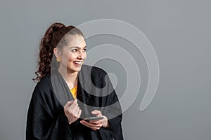 Curly-haired emotional teenager woman in a black cloak using mobile phone, isolated on gray background