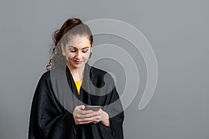 Curly-haired emotional teenager woman in a black cloak using mobile phone, isolated on gray background