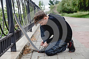 A curly-haired European teen in a black hoodie with skate sitting on the embankment and tying his shoelaces on sneakers