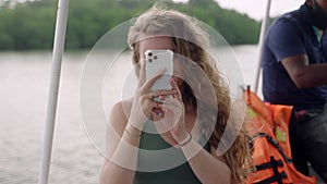 Curly-haired female moments on smartphone during river cruise, enjoys calm waters. Smiling, engaging with digital tech