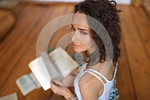 A curly-haired girl with books is sitting on the wooden floor.