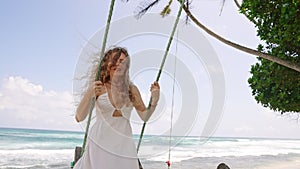 Curly-haired girl in white dress enjoys swing ride by turquoise sea. Breezy summer day, hair waves in wind, playful