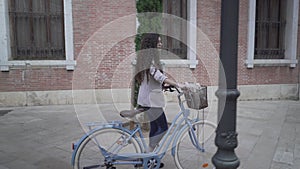 Curly-Haired Lady With Bicycle Is Walking Along The Sidestreet Of City Park. Tracking Shot