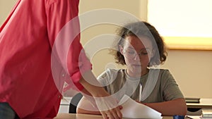 A curly-haired schoolgirl with glasses is writing a test and peeping at a cheat sheet. The girl laughs while sitting at