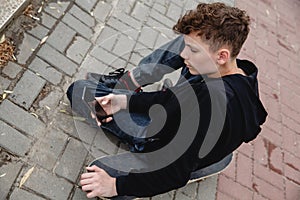 A curly-haired teen of European appearance in black hoodie sitting on an alley on a skateboard and talking on the phone