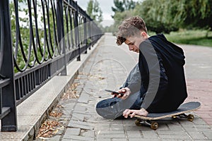 A curly-haired teen of European appearance in black hoodie sitting on an alley on a skateboard and talking on the phone