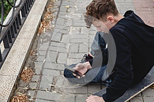 A curly-haired teen of European appearance in black hoodie sitting on an alley on a skateboard and talking on the phone
