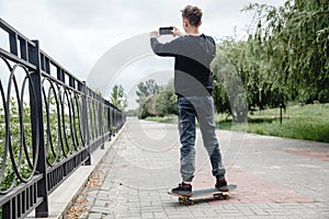 A curly-haired teenager of European appearance in a black hoodie standing on a skateboard in an alley takes a selfie