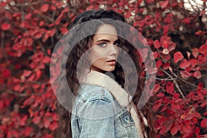 Curly haired Woman autumn outdoor portrait. Young beautiful brunette in jeans jacket with fur collar posing in red leaves park. A