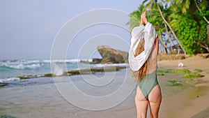 Curly-haired woman in green bikini walks on sandy beach, waves white shawl by clear sea. Invites viewers for tropical